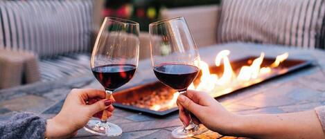 Dos personas brindando con copas de vino en una mesa al aire libre junto a una chimenea.