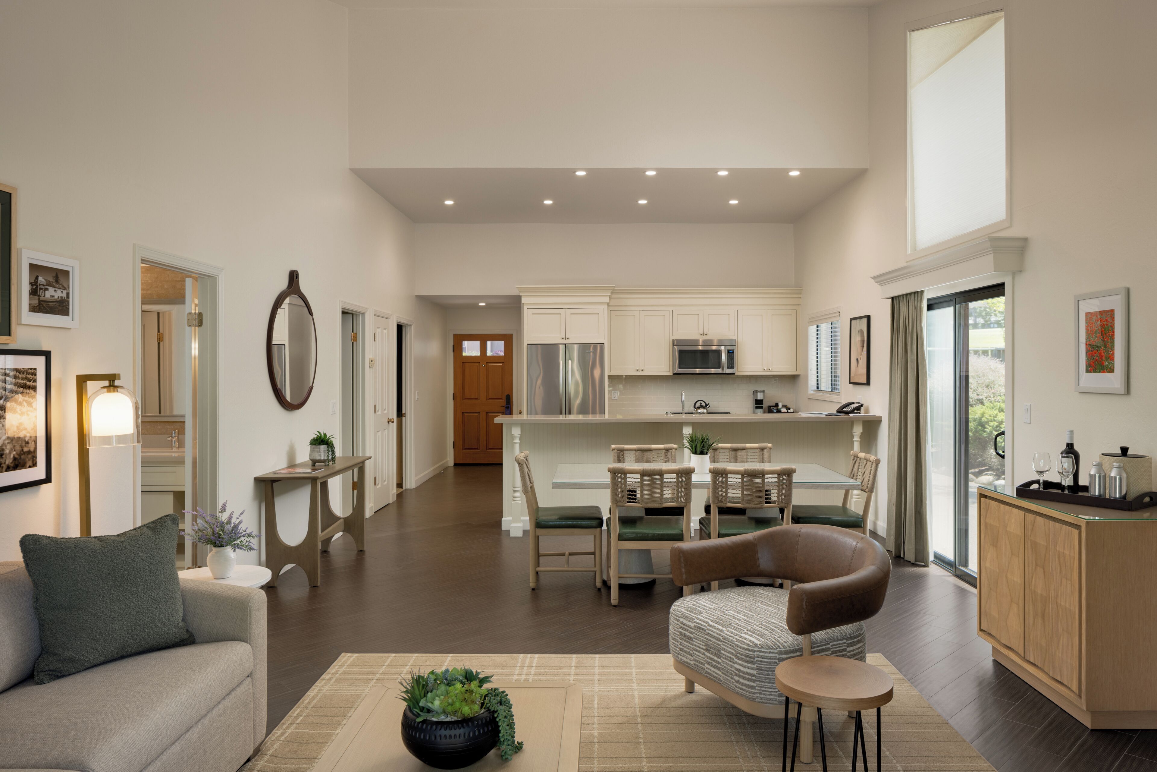 A spacious living area with a sofa, a coffee table, and a dining table with chairs. A kitchen with a refrigerator and microwave is visible in the background.