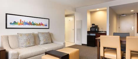 A hotel room with a sofa, ottoman, and chairs, a dining table, a kitchen area, and a framed cityscape picture on the wall.