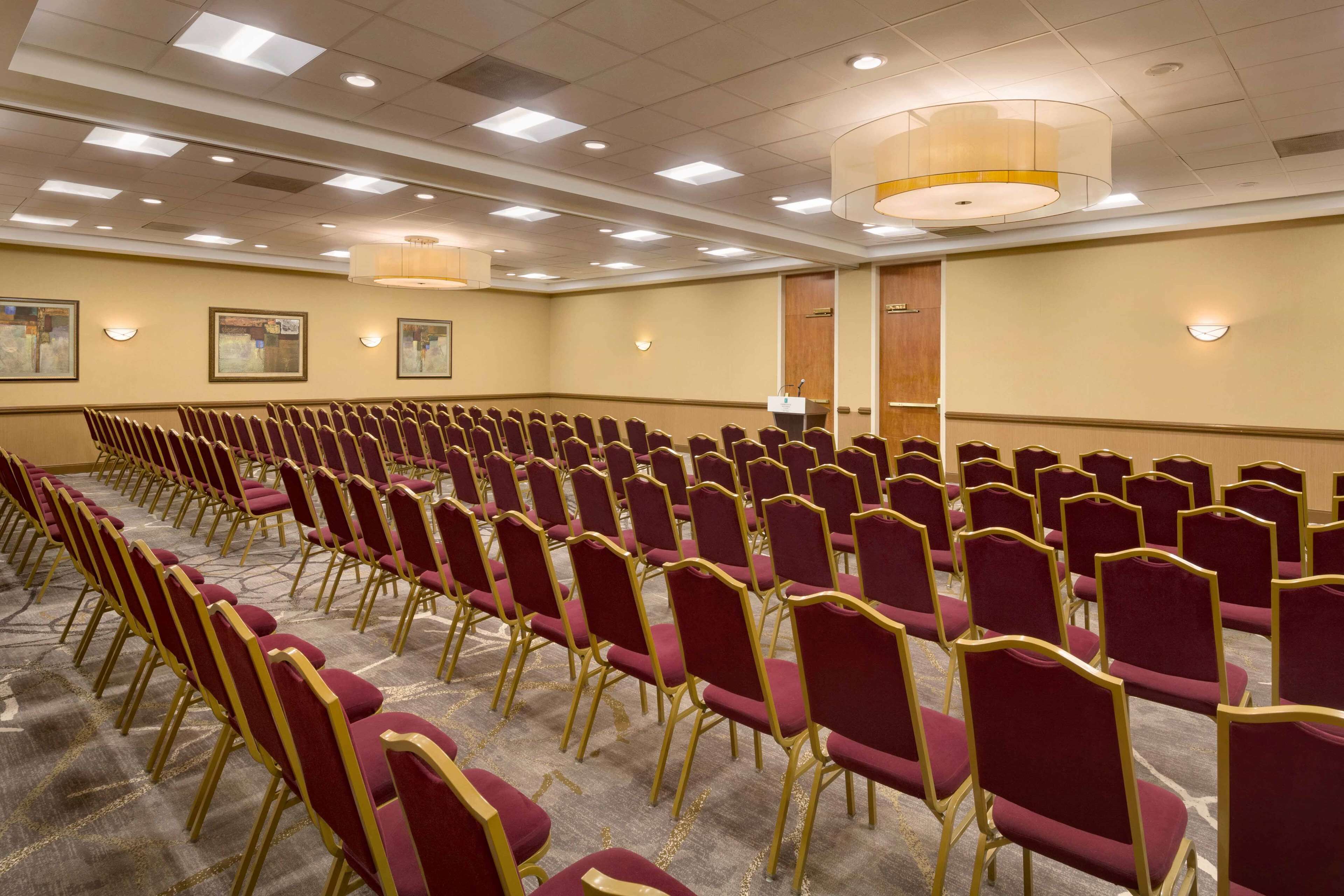 A conference room with rows of red chairs and gold frames, carpeted floor, and recessed lighting.