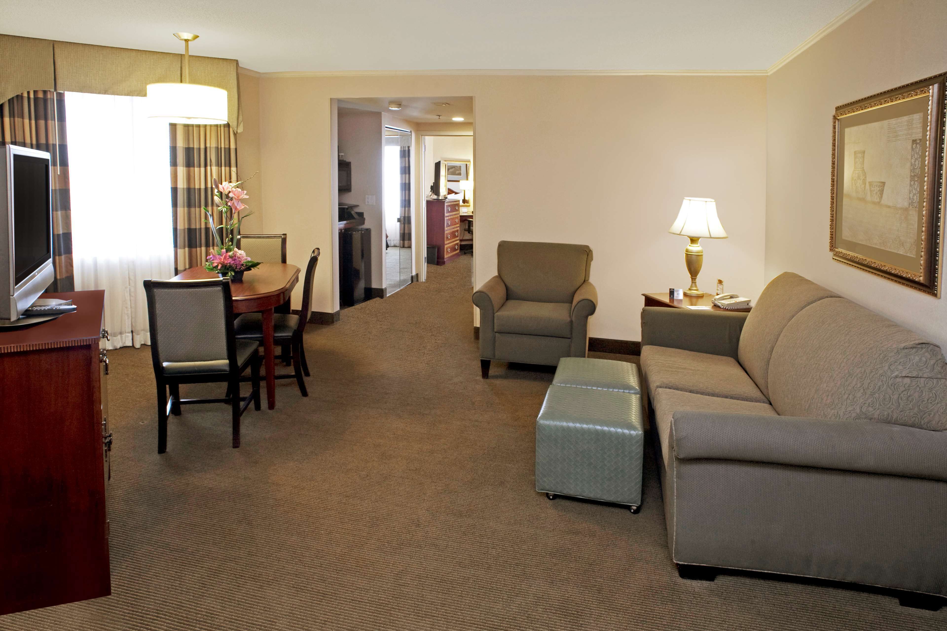 A hotel room with a TV, a dining table with chairs, a sofa, an ottoman, a lamp, and a framed picture on the wall.