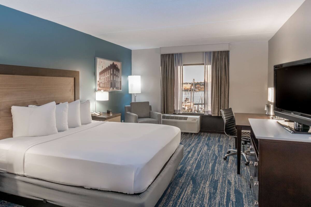 Habitación de hotel con una cama grande, un escritorio con televisor, una silla, una lámpara y una ventana con vistas al puerto.