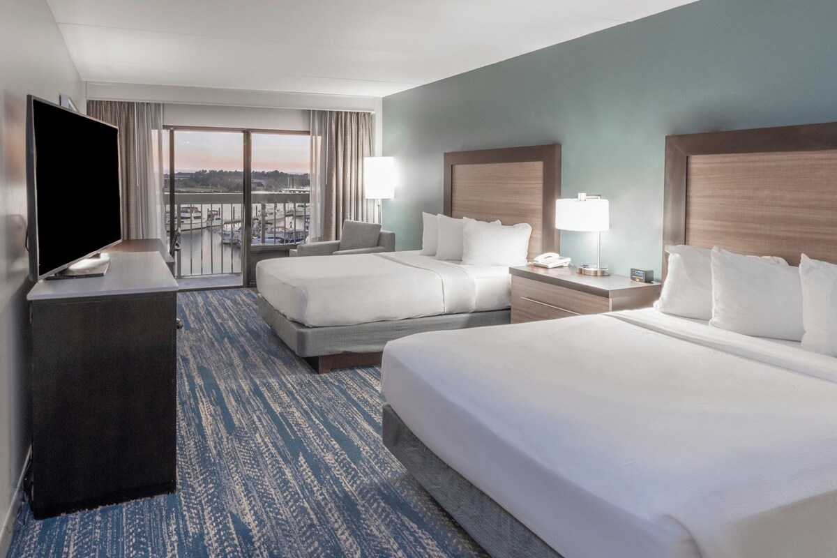 A hotel room with two beds, a TV, a desk, and a view of the city.