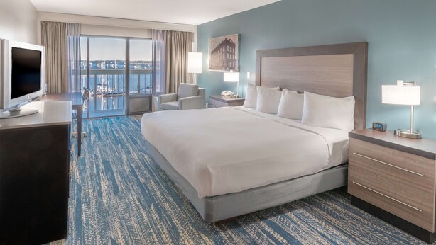 A hotel room with a large bed, a desk with a TV, a chair, a nightstand with a lamp, and a view of the city through large windows. - DoubleTree by Hilton New Bern Riverfront (New Bern)