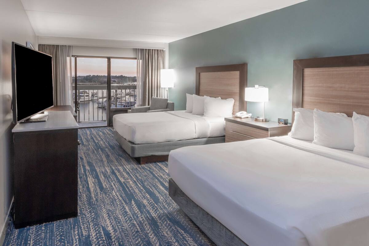 Habitación de hotel con dos camas, televisión, escritorio y balcón con vista a la ciudad.