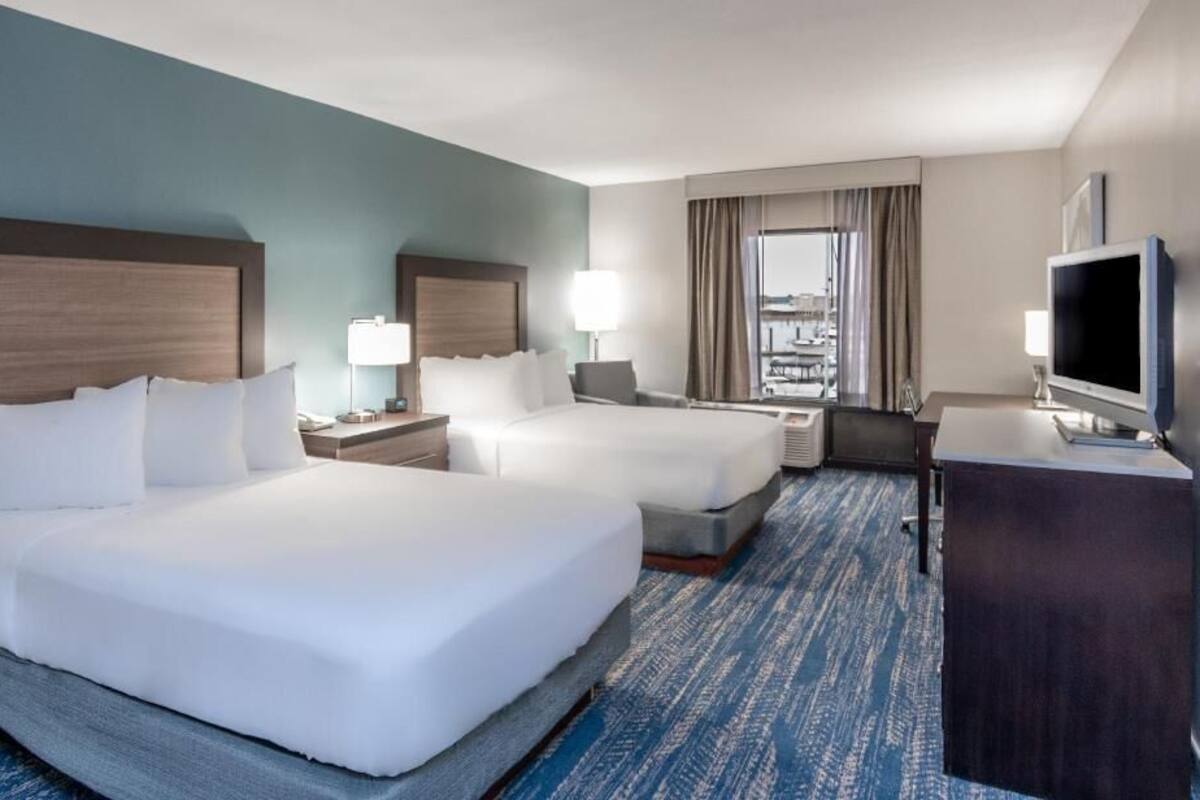 A hotel room with two beds, a desk, a TV, and a view of the city.