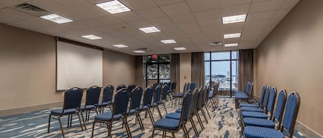 Un salón de conferencias con filas de sillas, una pantalla de proyector y un amplio ventanal con vistas a la ciudad.
