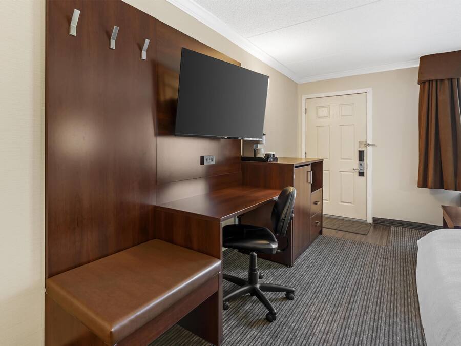 Ett hotellrum med ett skrivbord i trä, en tv som hänger ovanför skrivbordet, en svart kontorsstol, en säng och en dörr.