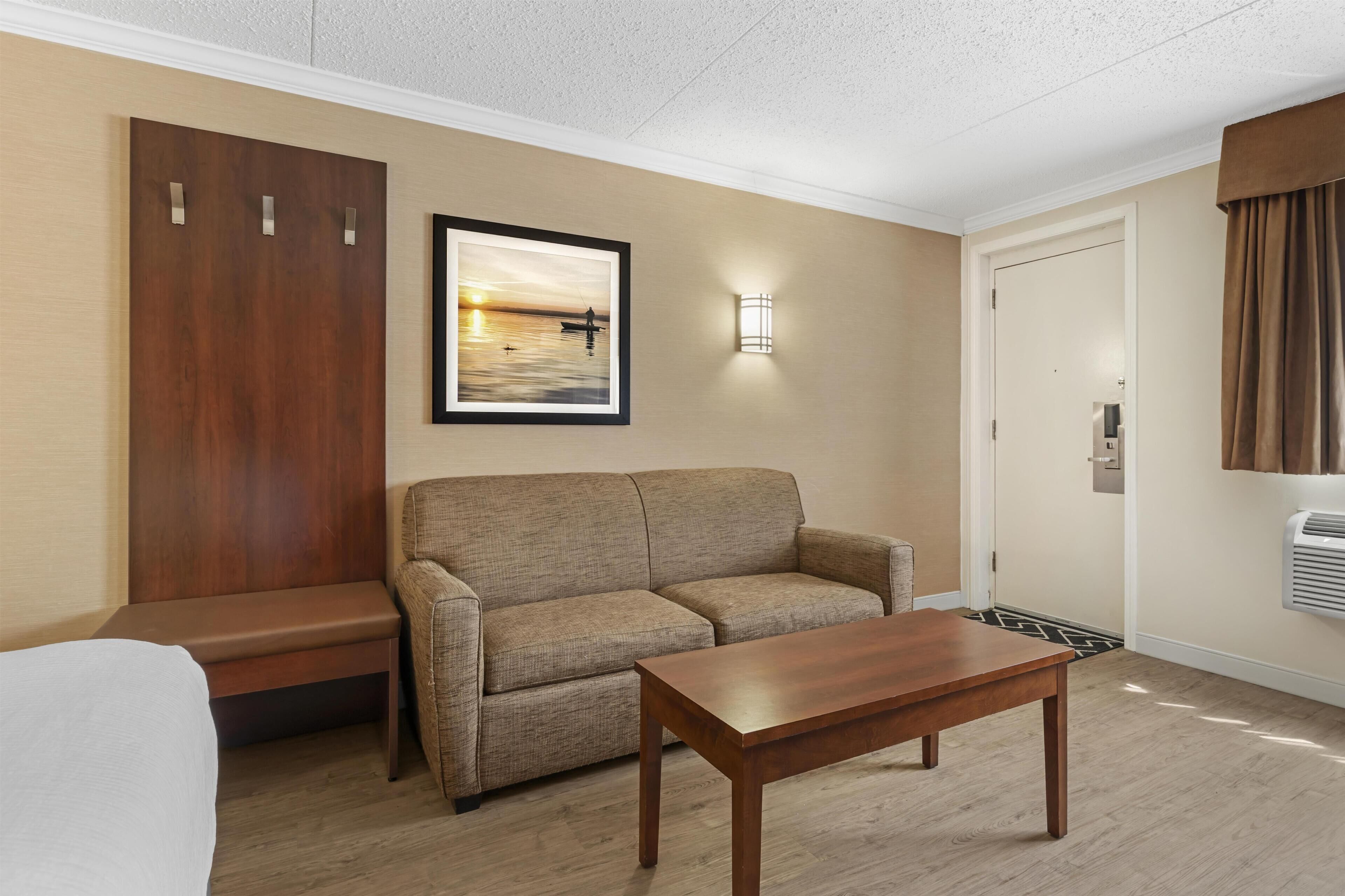 Une chambre d’hôtel avec un canapé, une table en bois, un banc, un tableau encadré, un luminaire fixé au mur et une porte.