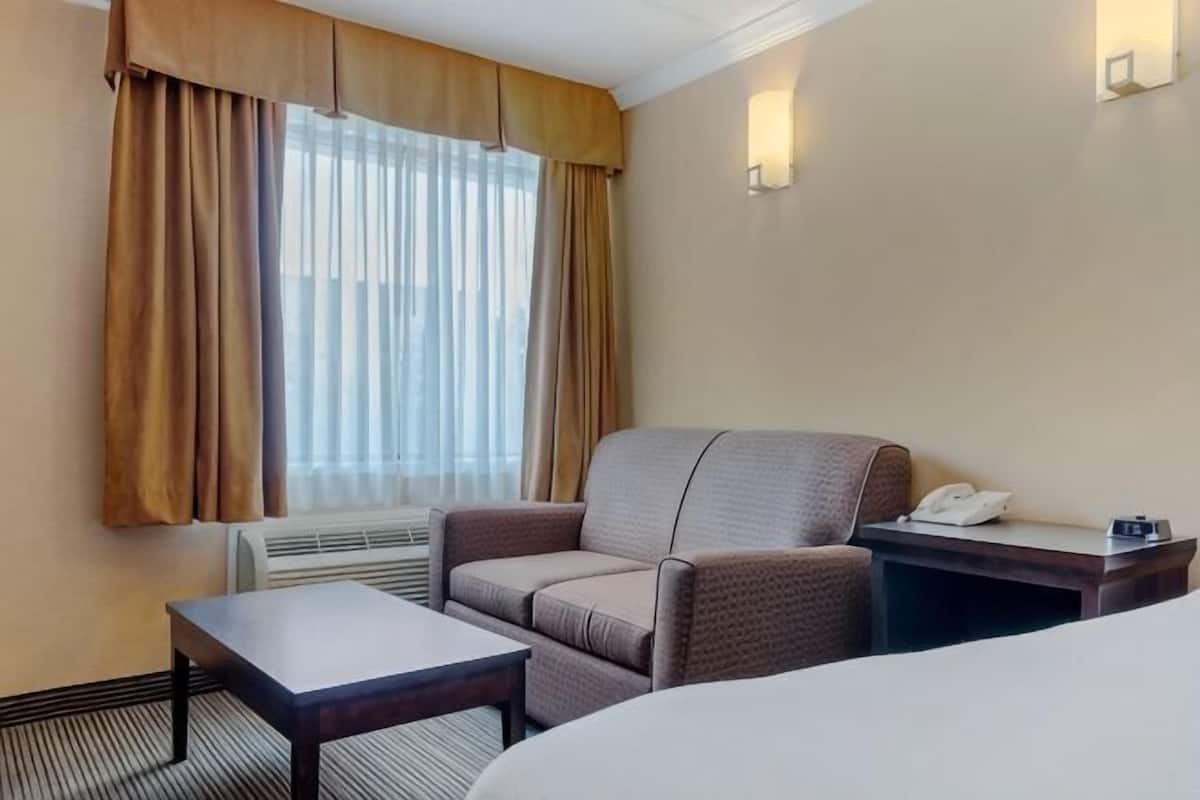 A hotel room with a bed, a chair, a small table, a bedside table with a phone, and a window with curtains.