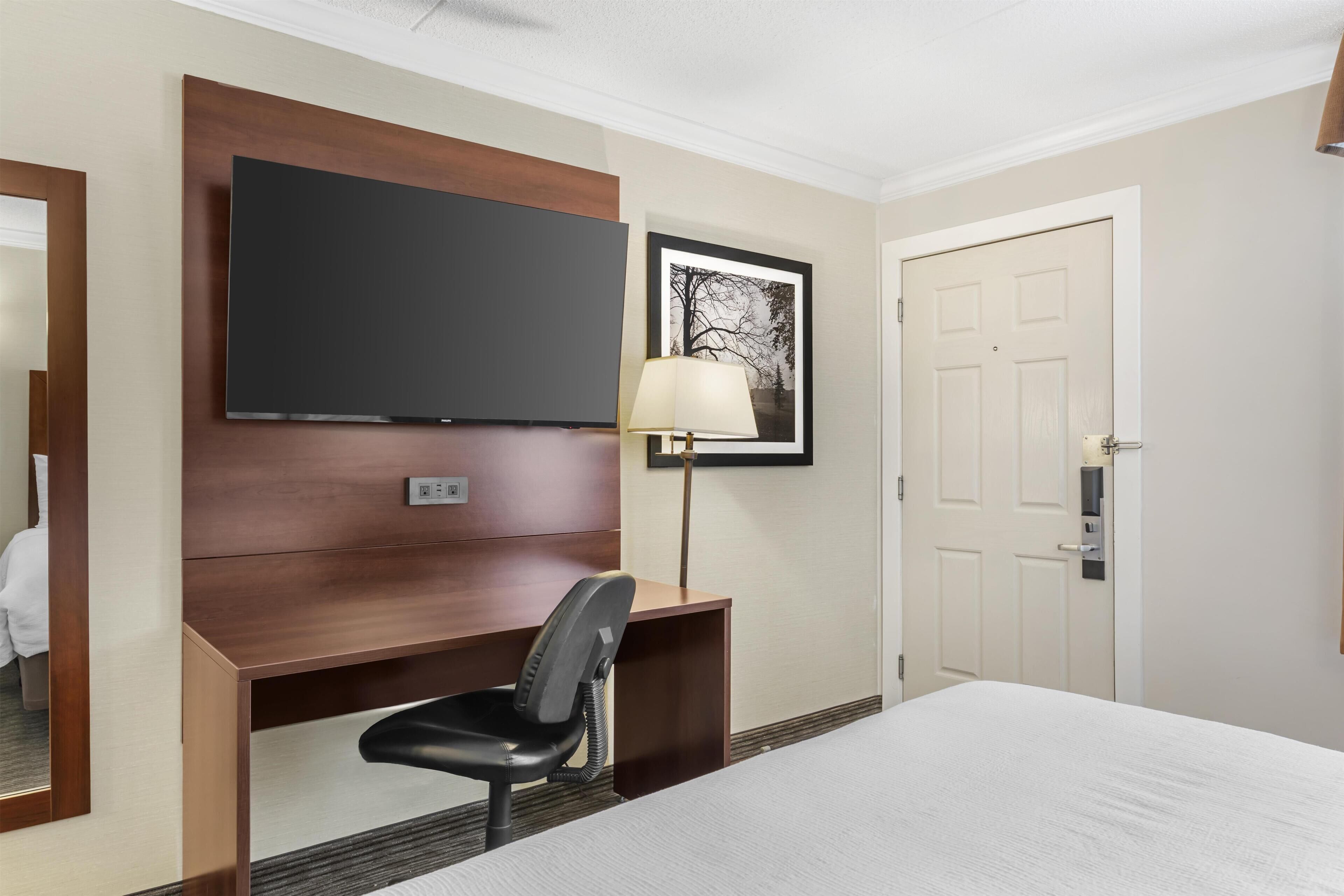 A hotel room with a bed, a desk with a chair, a television, a framed picture, and a door.