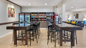A modern indoor dining area with a glass-top table, black bar stools, and shelves stocked with snacks and beverages. - Comfort Inn & Suites Danbury-Bethel (Danbury)