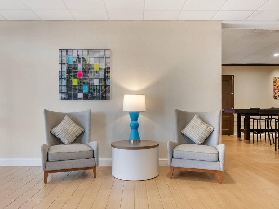 A modern living room with two grey armchairs, a round side table with a blue lamp, and a cityscape artwork on the wall.