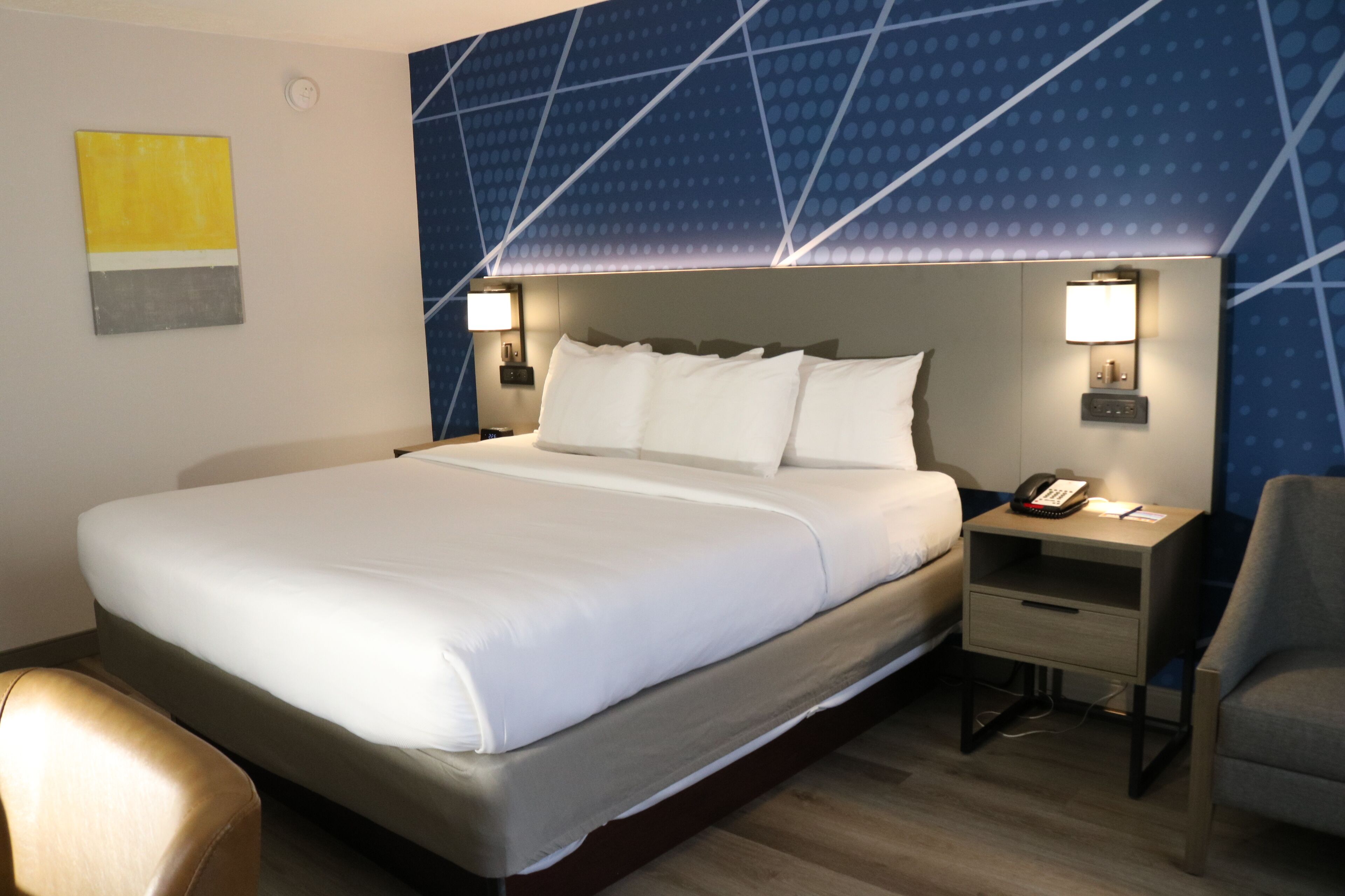 A hotel room with a large bed, two bedside tables with lamps, a chair, and a wall with a geometric pattern.