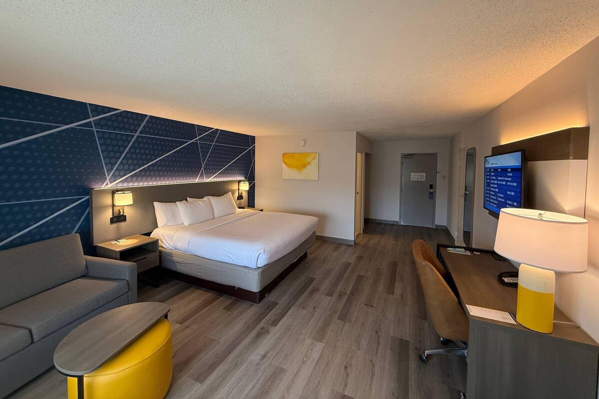 A hotel room with a bed, a desk, a chair, a sofa, a TV, and a wall-mounted lamp.