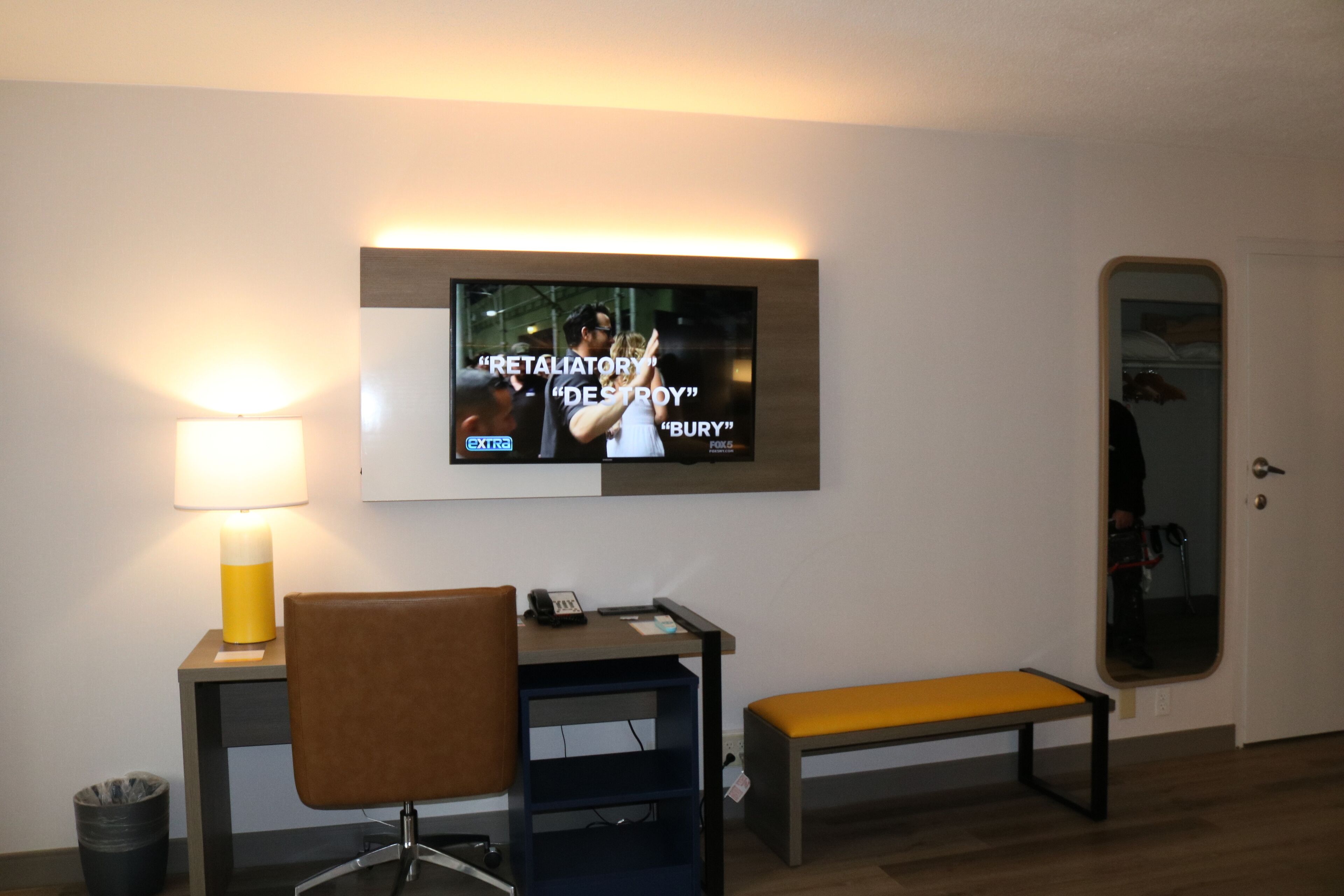 A hotel room with a TV mounted on the wall displaying a movie scene, a desk with a chair, a bench, and a lamp.