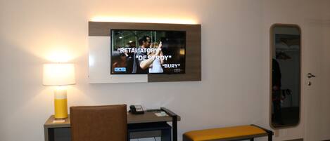 A hotel room with a TV mounted on the wall displaying a movie scene, a desk with a chair, a bench, and a lamp.