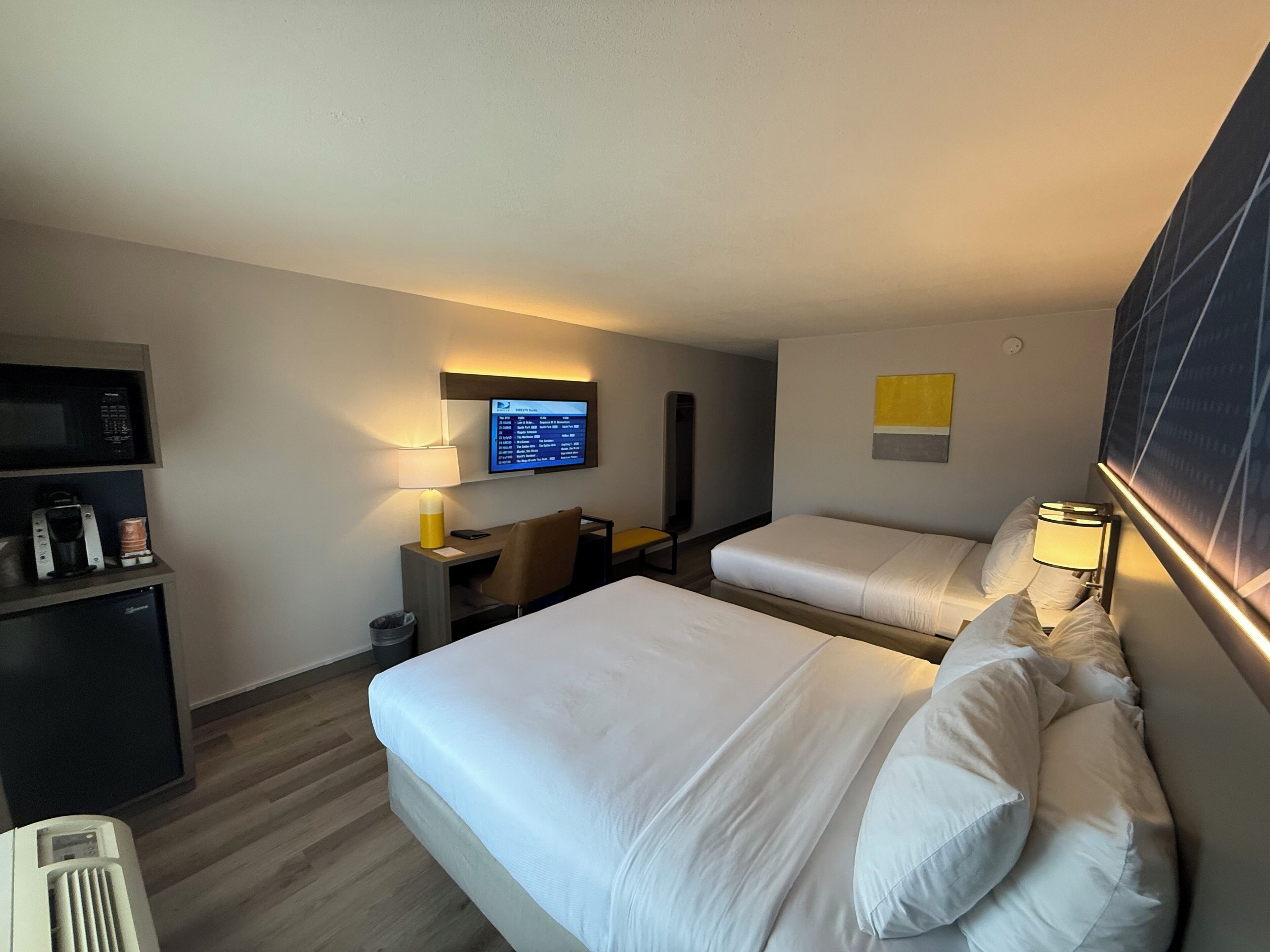 A hotel room with two beds, a desk with a computer, a TV mounted on the wall, and a small seating area.