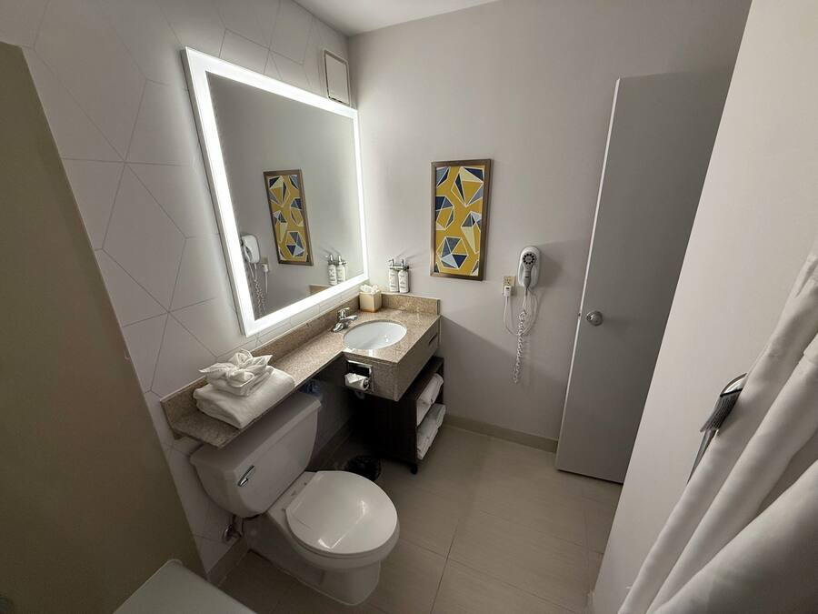 Un baño con inodoro, lavamanos y espejo. Hay toallas en el mostrador y una cortina de ducha parcialmente visible.