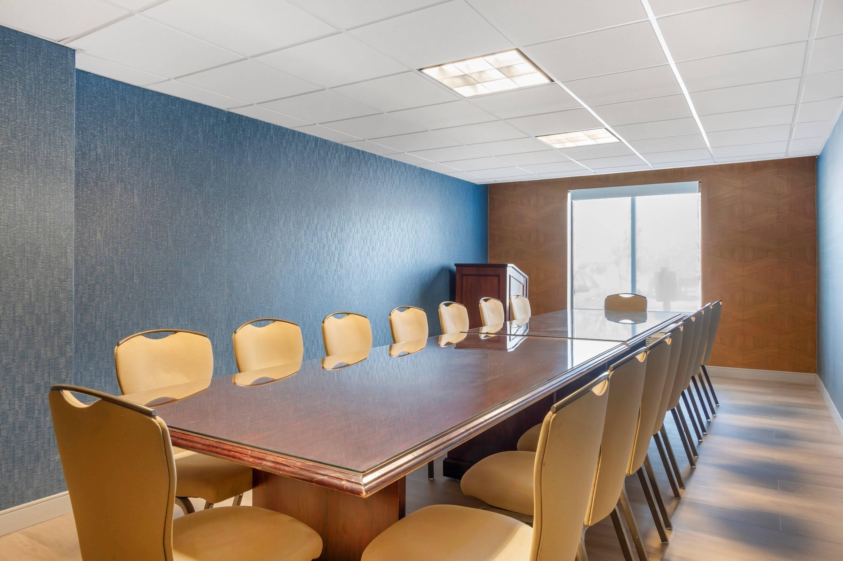 A conference room with a long table, beige chairs, a wooden cabinet, and a large window.