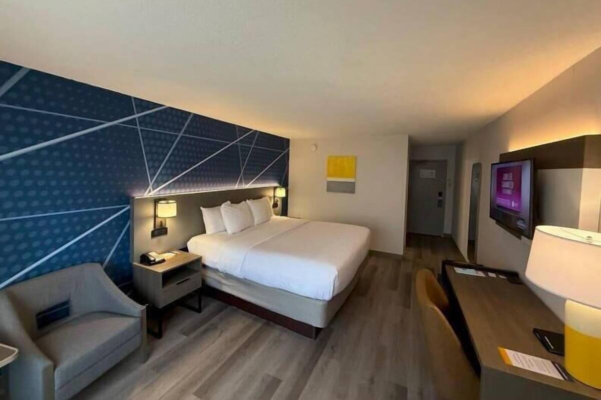 A hotel room with a bed, bedside tables, a chair, a desk, a TV, and a wall with geometric patterns.