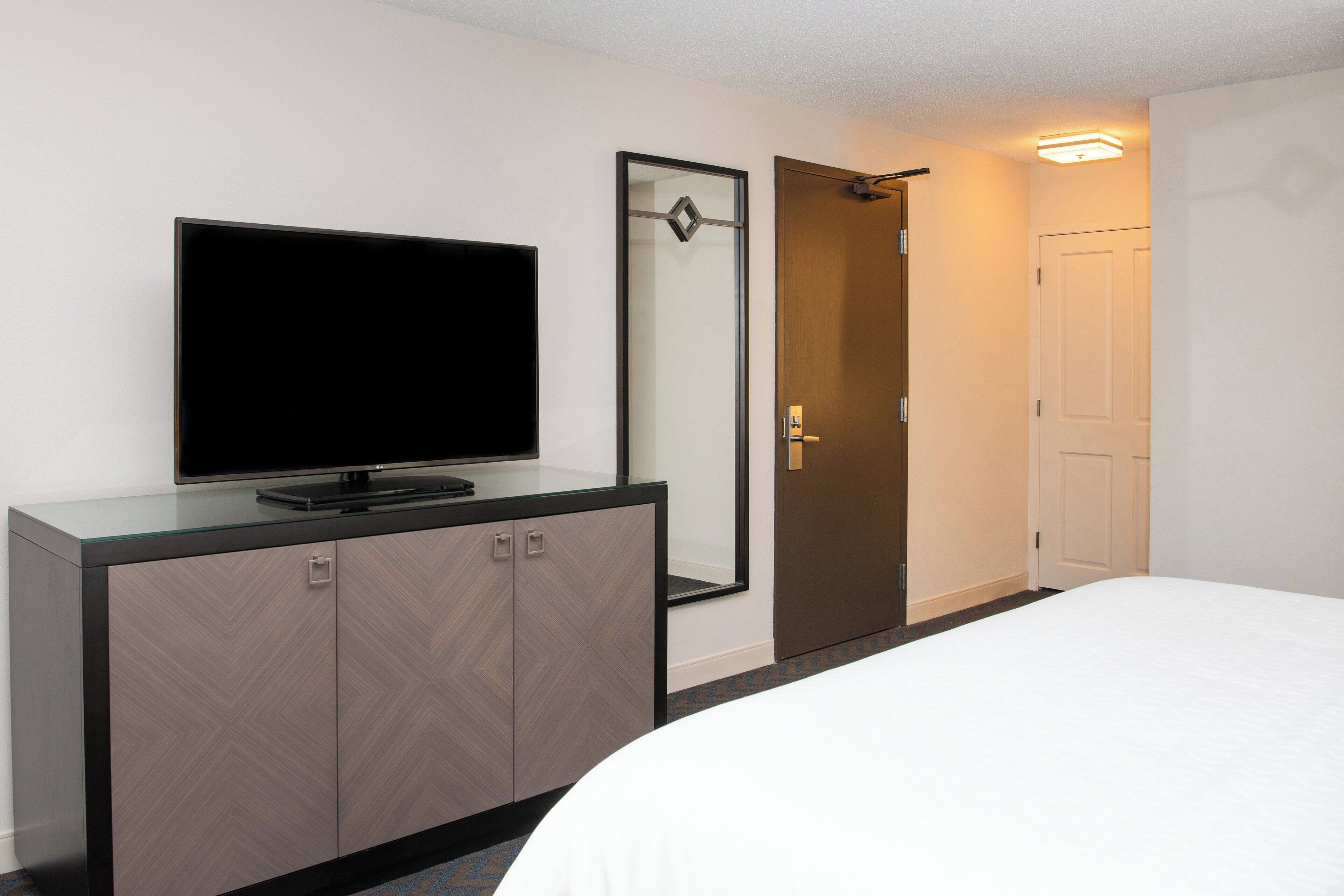 A hotel room with a bed, a TV on a cabinet, a mirror, and a door.