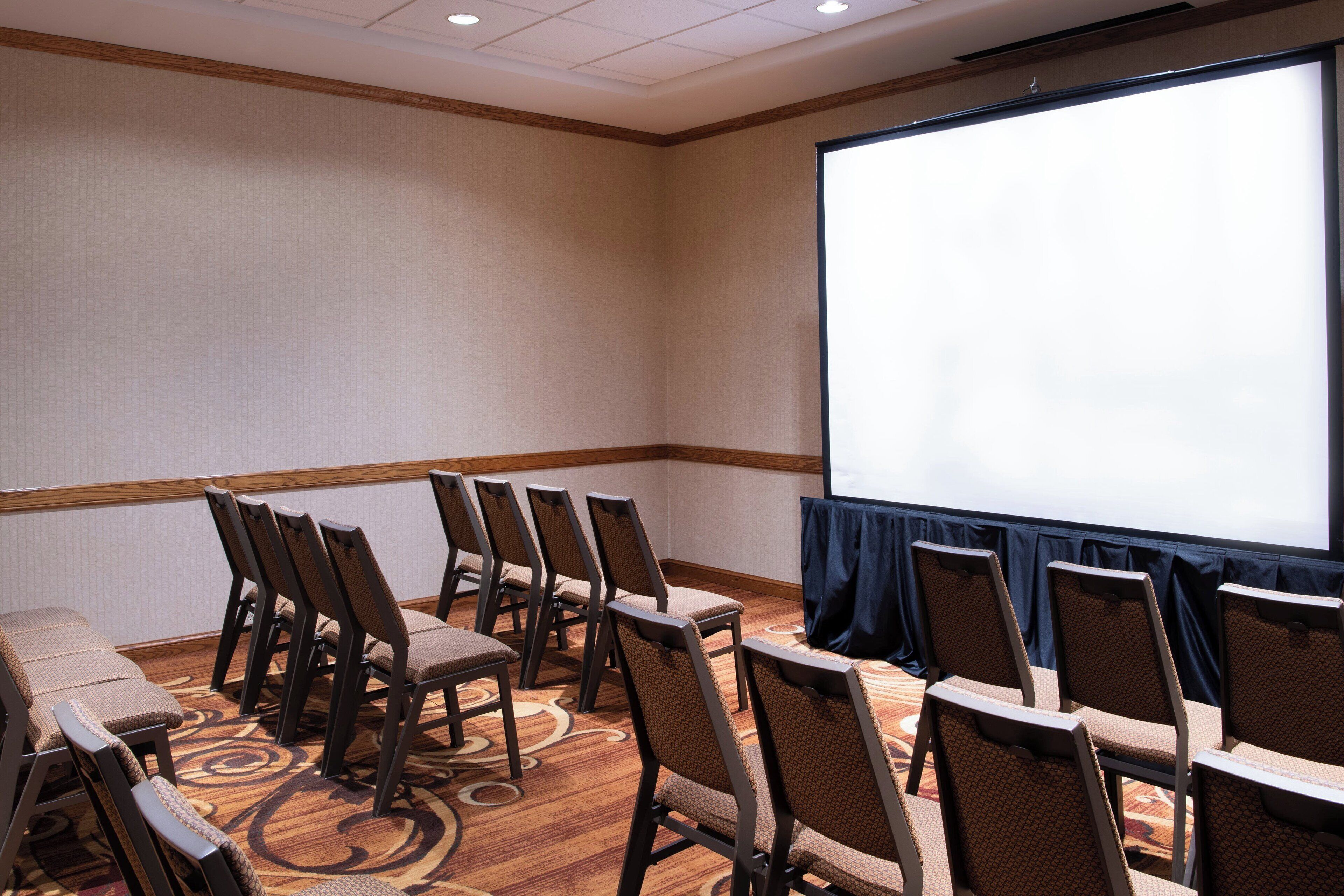 A conference room with rows of chairs, a large screen, and a carpeted floor.