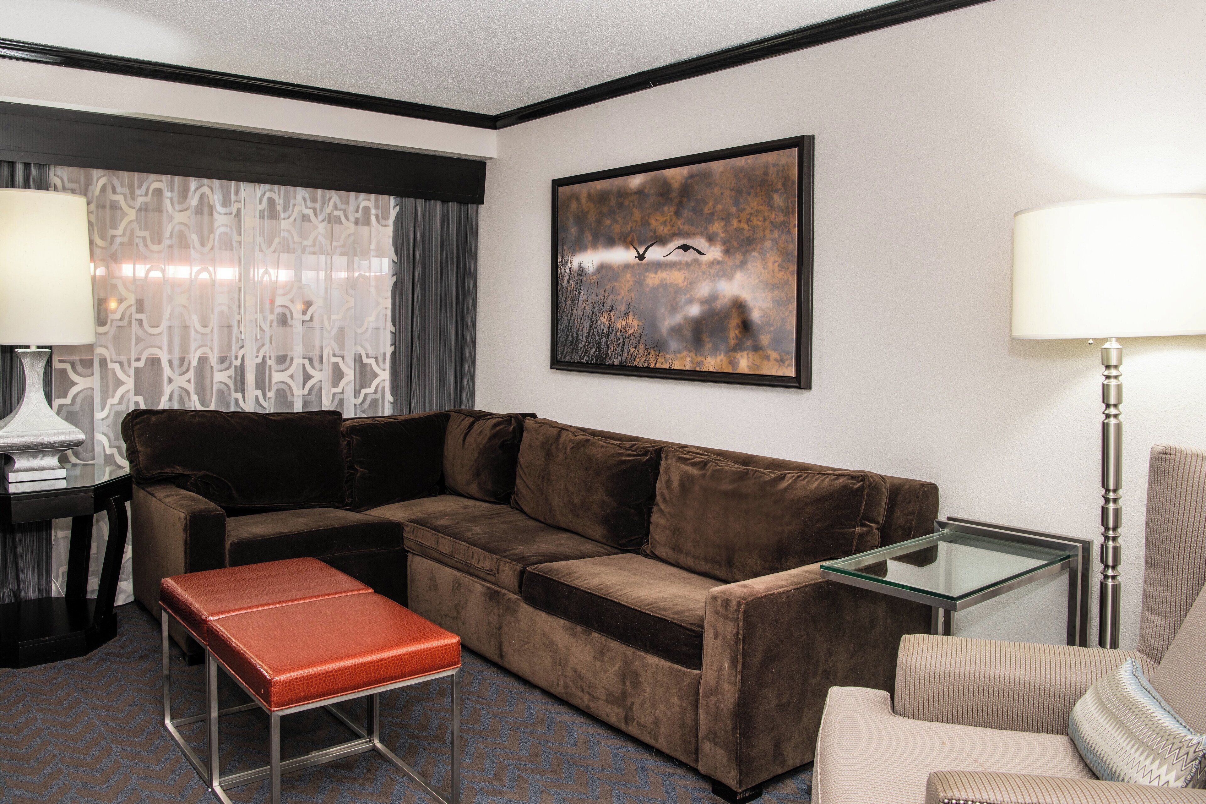 A hotel room with a brown sofa, a glass coffee table, a red bench, a patterned curtain, and a framed artwork on the wall.
