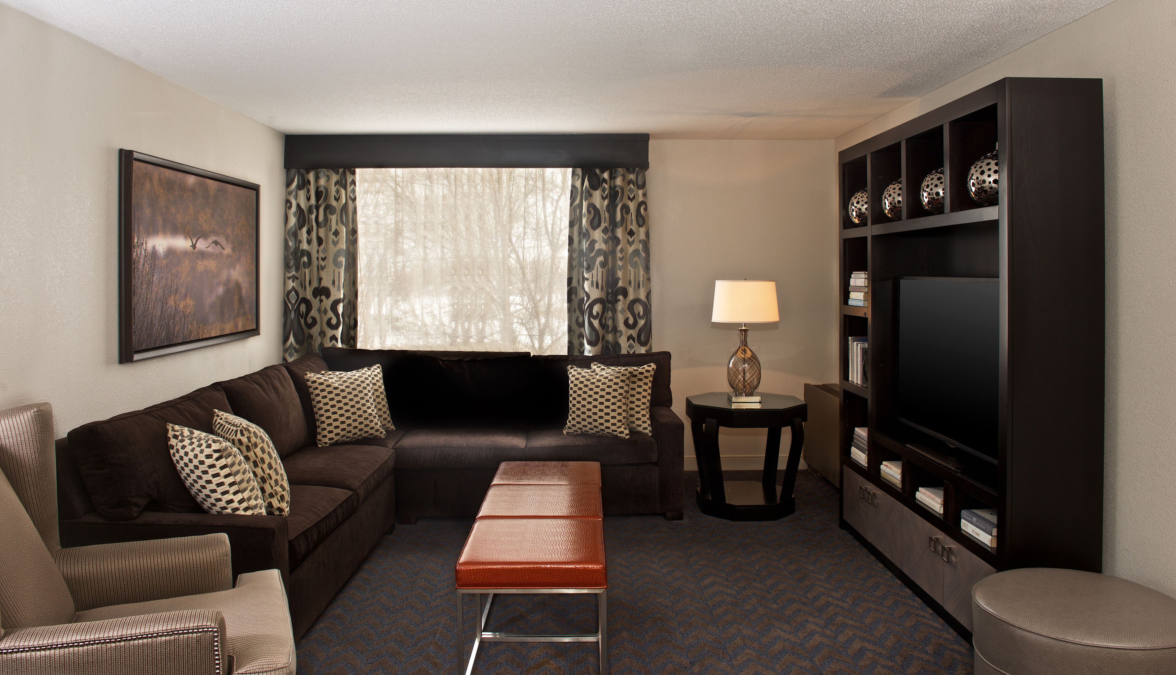 A hotel room with a dark brown sofa, patterned cushions, a small wooden table with a lamp, a television on a stand, and a bookshelf.
