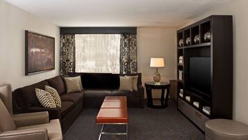A hotel room with a dark brown sofa, patterned cushions, a small wooden table with a lamp, a television on a stand, and a bookshelf.
