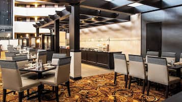 A restaurant with a modern interior, featuring a patterned carpet, dark wooden furniture, and a view of the kitchen in the background.