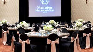 A Sheraton Minneapolis West hotel conference room with round tables, black chairs with peach sashes, white floral centerpieces, and a large screen displaying the hotel logo.