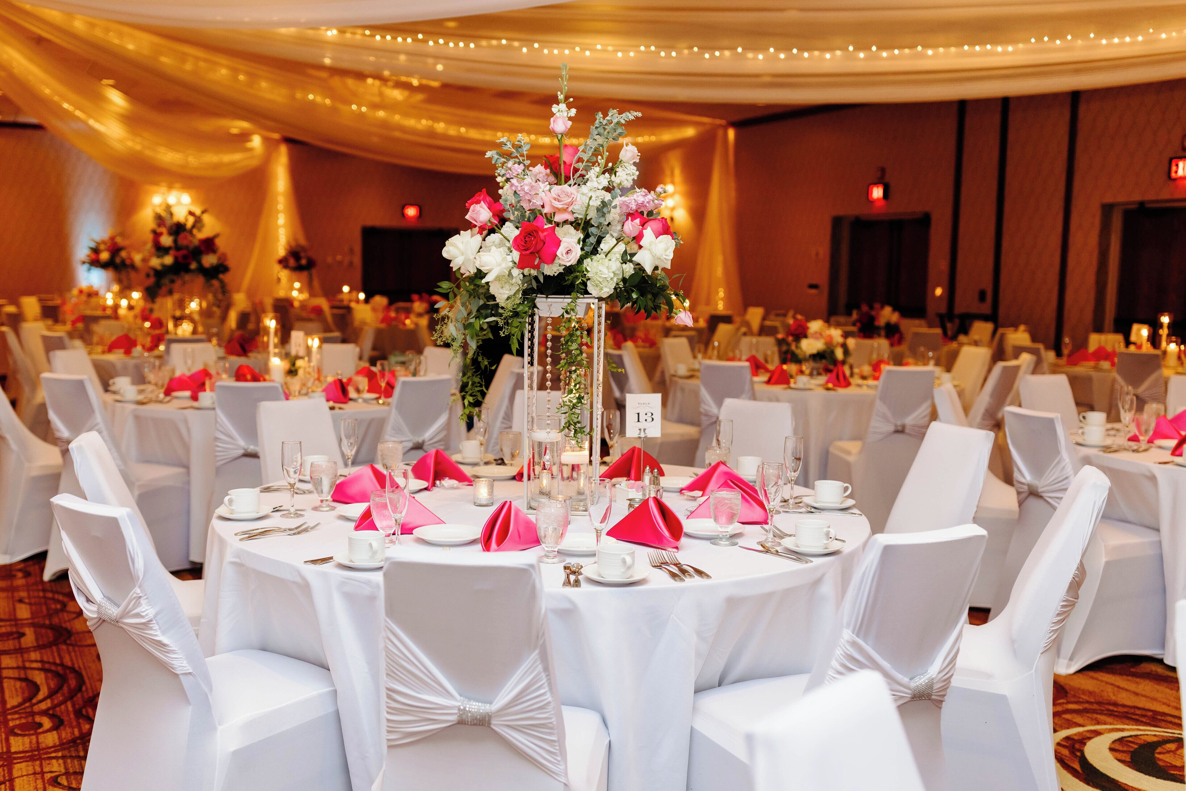 A banquet hall with round tables, white chairs, and a central floral arrangement. Tables are set with pink napkins and white tablecloths.