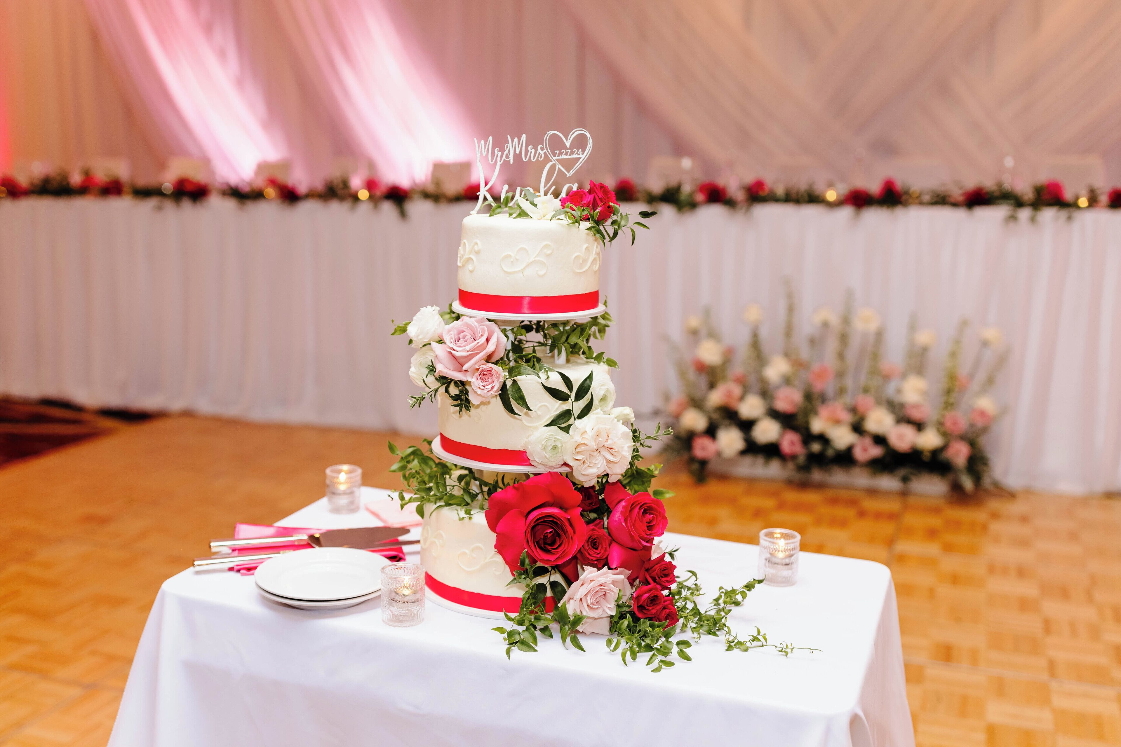 Um bolo de casamento de vários andares, com um enfeite no topo, rodeado de rosas, sobre uma mesa com velas e talheres.
