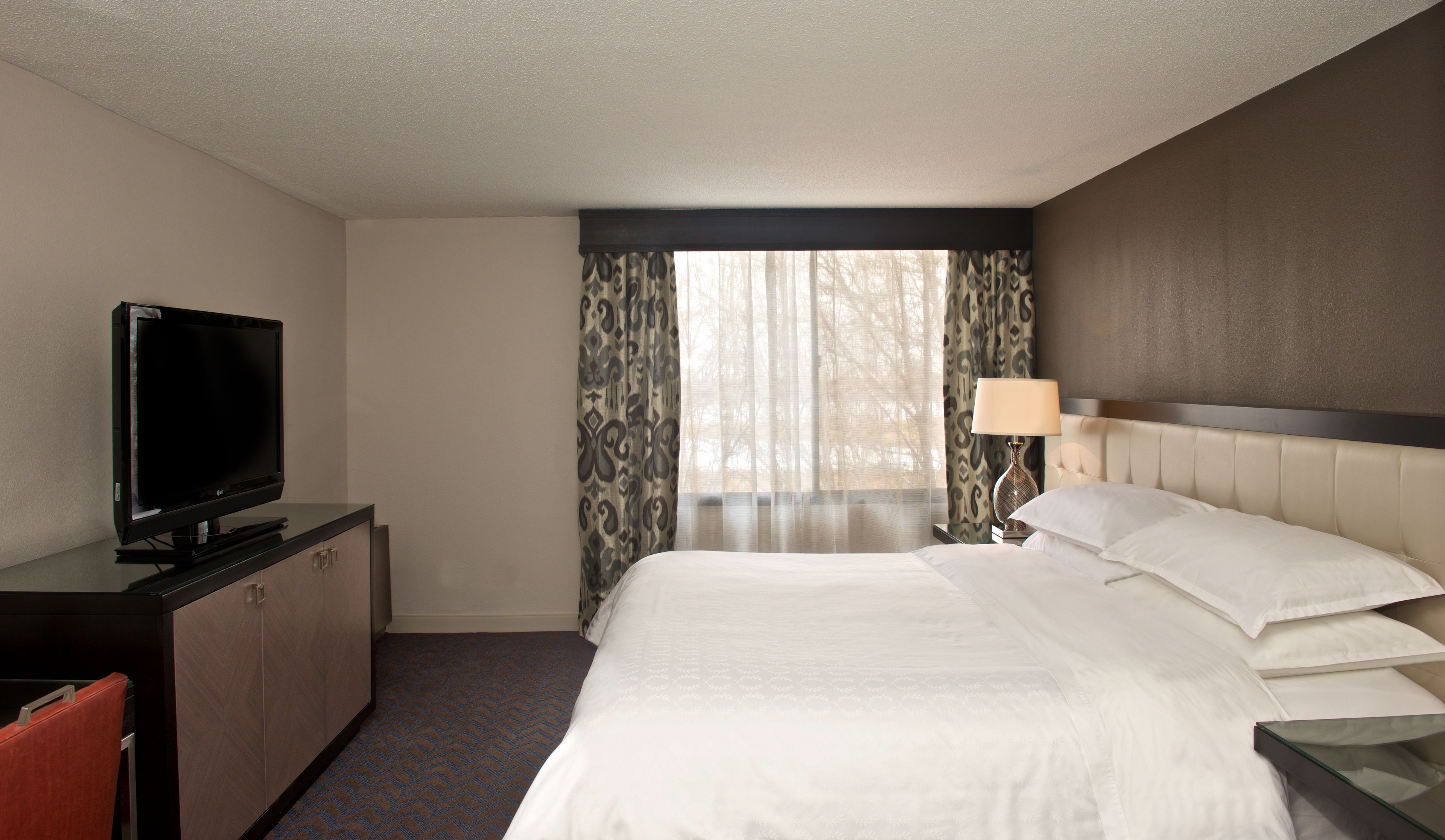 A hotel room with a bed, a television on a cabinet, a window with curtains, and a bedside lamp.