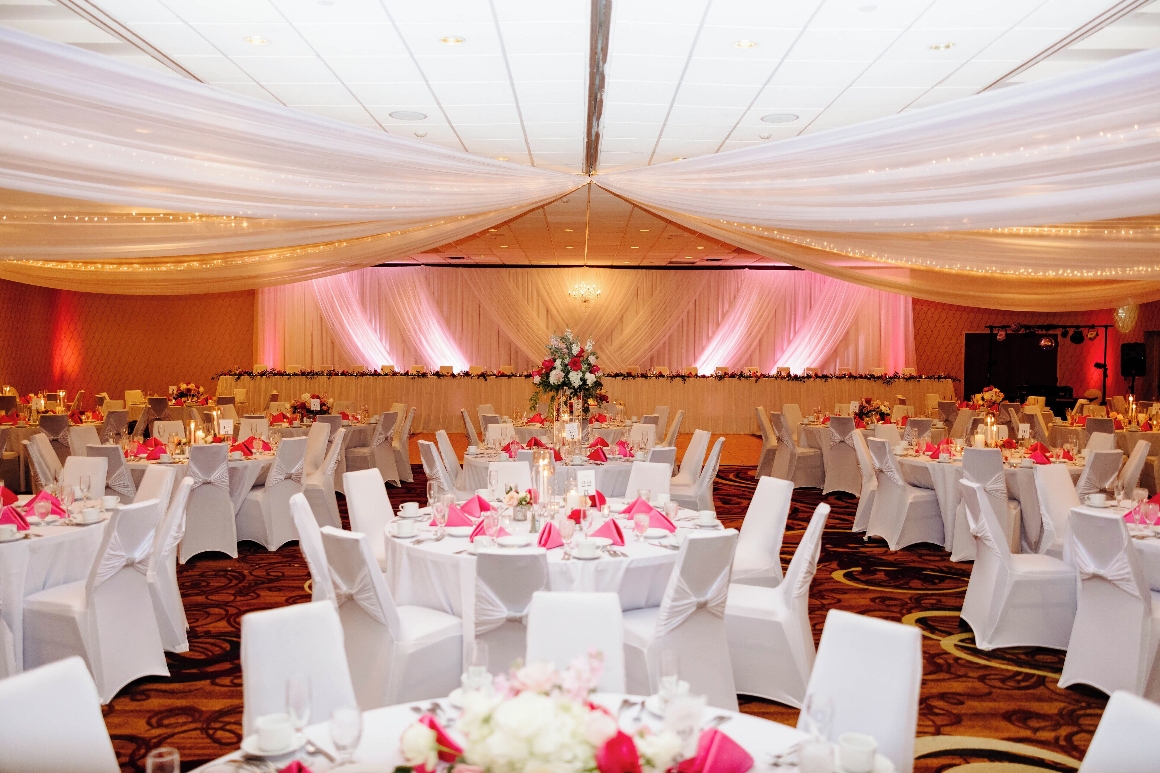 A banquet hall with round tables, white chairs, and draped ceiling. Tables are set with pink napkins and white tablecloths.