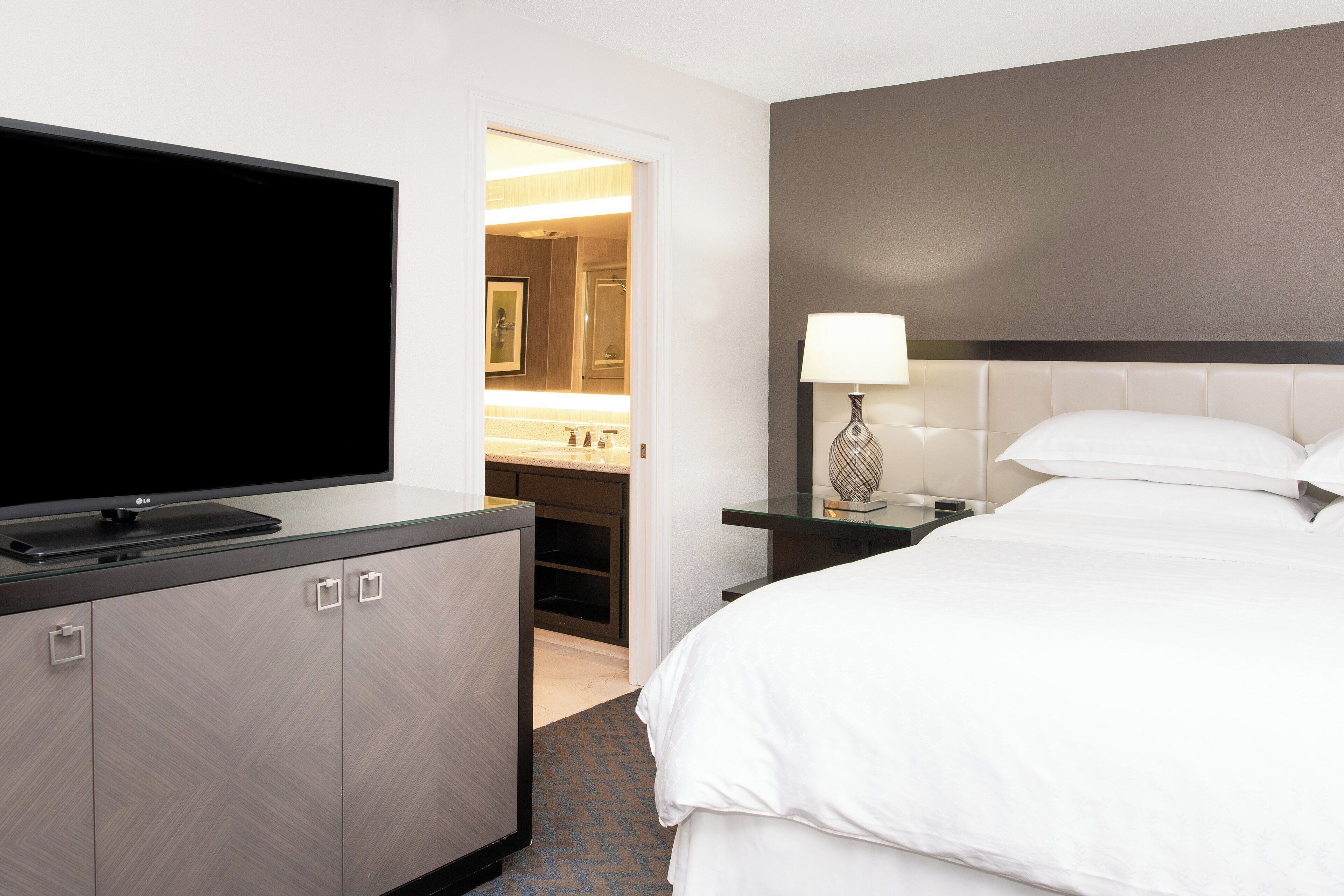 Habitación de hotel con una televisión de pantalla plana sobre un escritorio con cubierta de vidrio, una cama con ropa blanca, una mesita de noche con lámpara, y un baño visible a través de una puerta abierta.