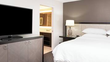 Habitación de hotel con una televisión de pantalla plana sobre un escritorio con cubierta de vidrio, una cama con ropa blanca, una mesita de noche con lámpara, y un baño visible a través de una puerta abierta.