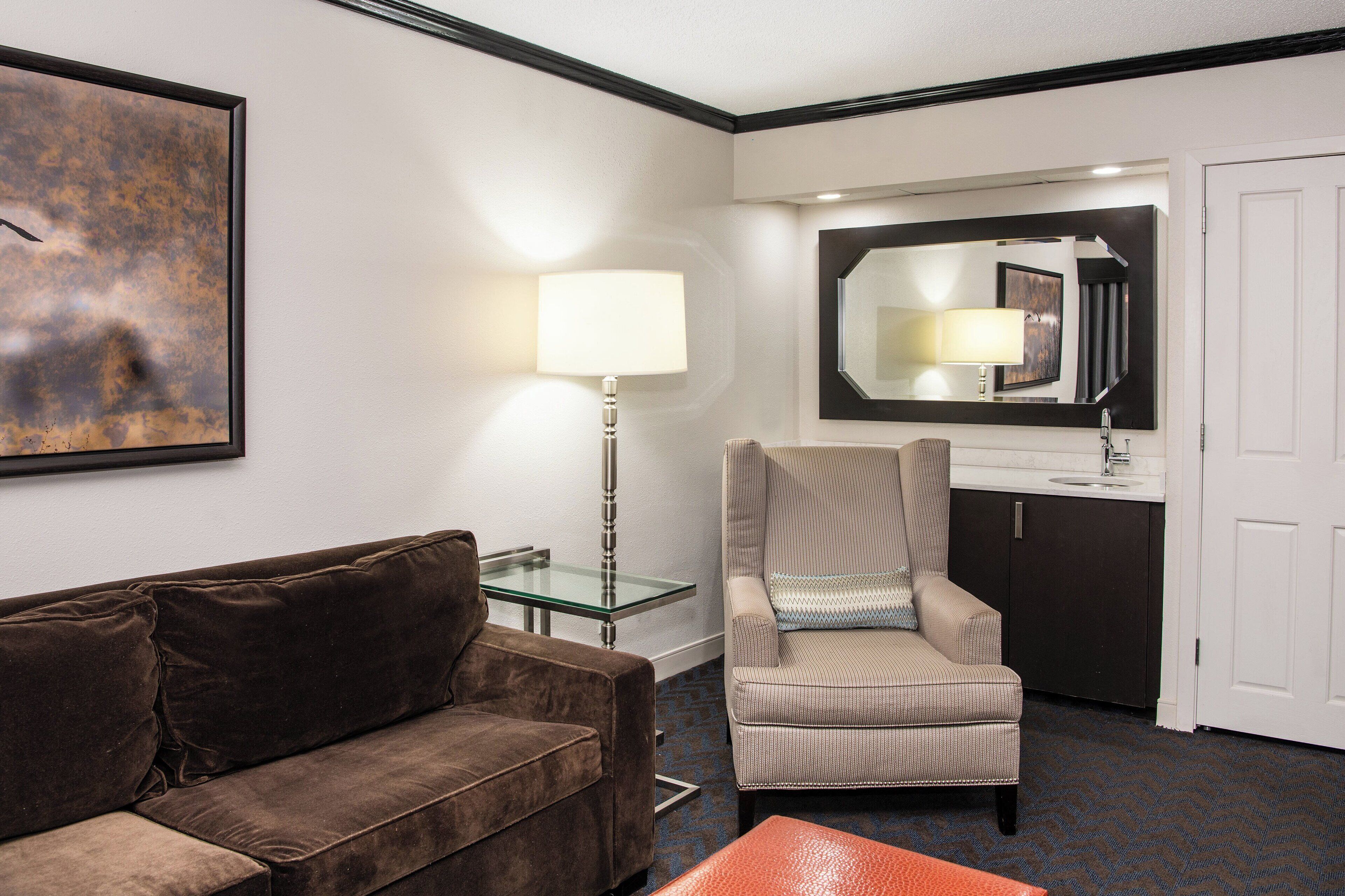 A hotel room with a brown sofa, a striped armchair, a glass table, a lamp, a mirror, a sink, and a door.