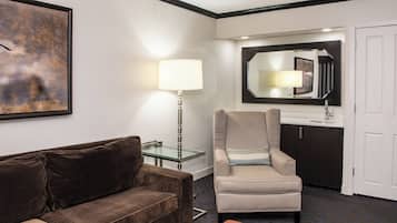 A hotel room with a brown sofa, a striped armchair, a glass table, a lamp, a mirror, a sink, and a door.