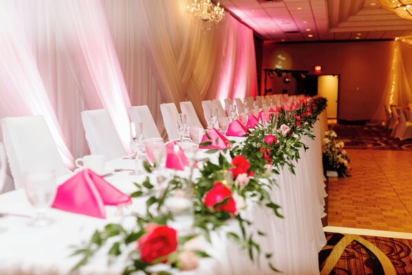Una mesa de banquete dispuesta con sillas blancas, servilletas rosas y centros de mesa florales.