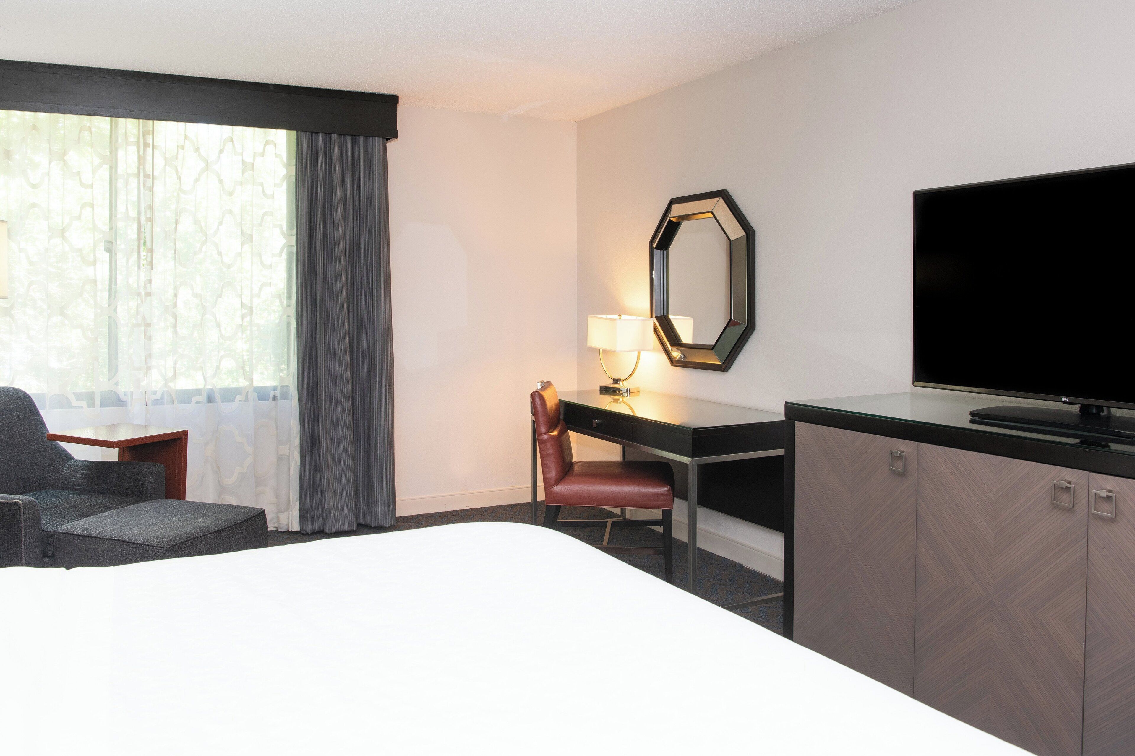 Una habitación de hotel con cama, escritorio, silla, televisor y ventana con cortinas.