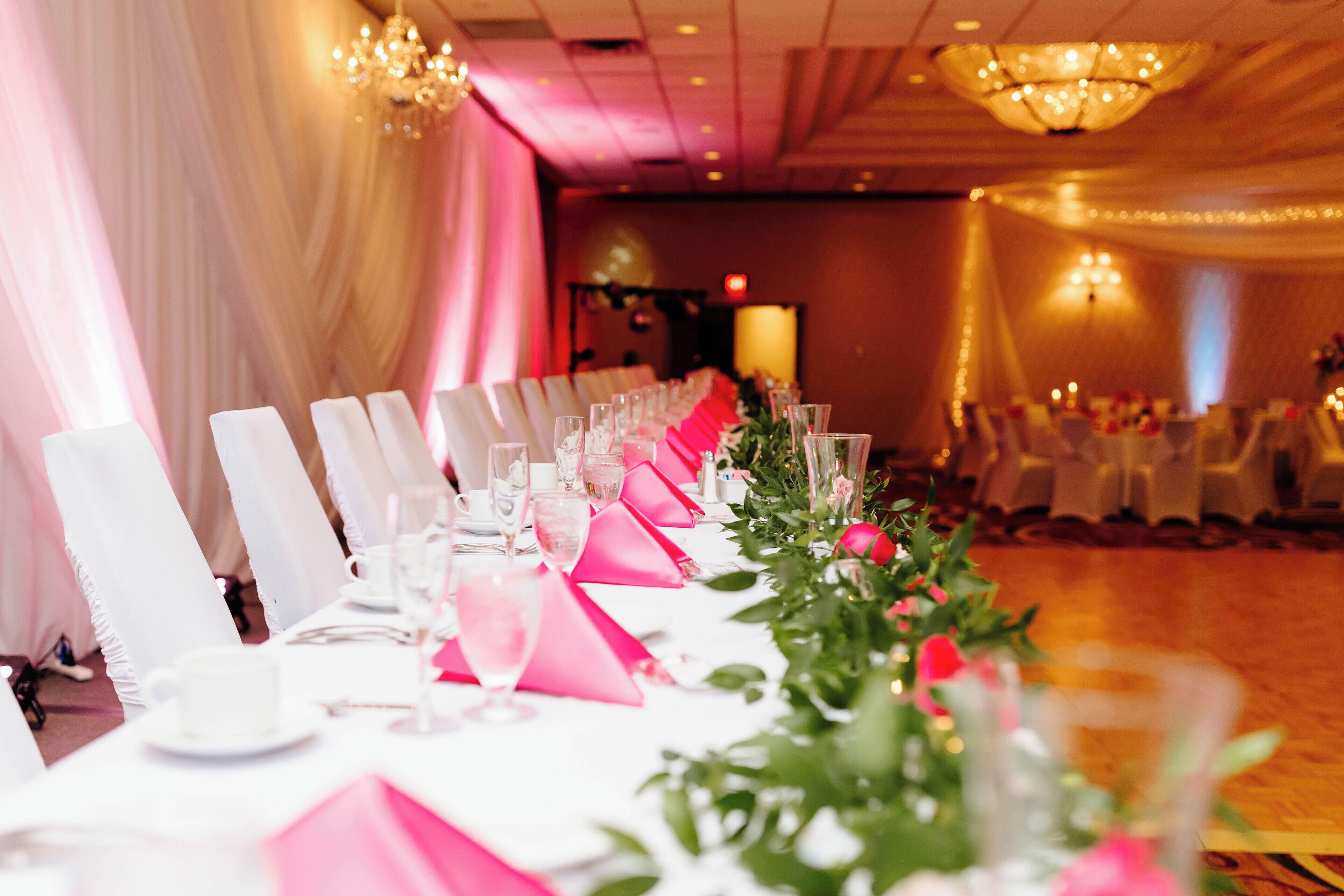 Une salle de banquet avec de longues tables dressées pour un événement formel, agrémentée de chaises blanches, de nappes roses et de centres de table floraux.