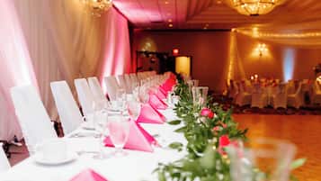 Une salle de banquet avec de longues tables dressées pour un événement formel, agrémentée de chaises blanches, de nappes roses et de centres de table floraux.
