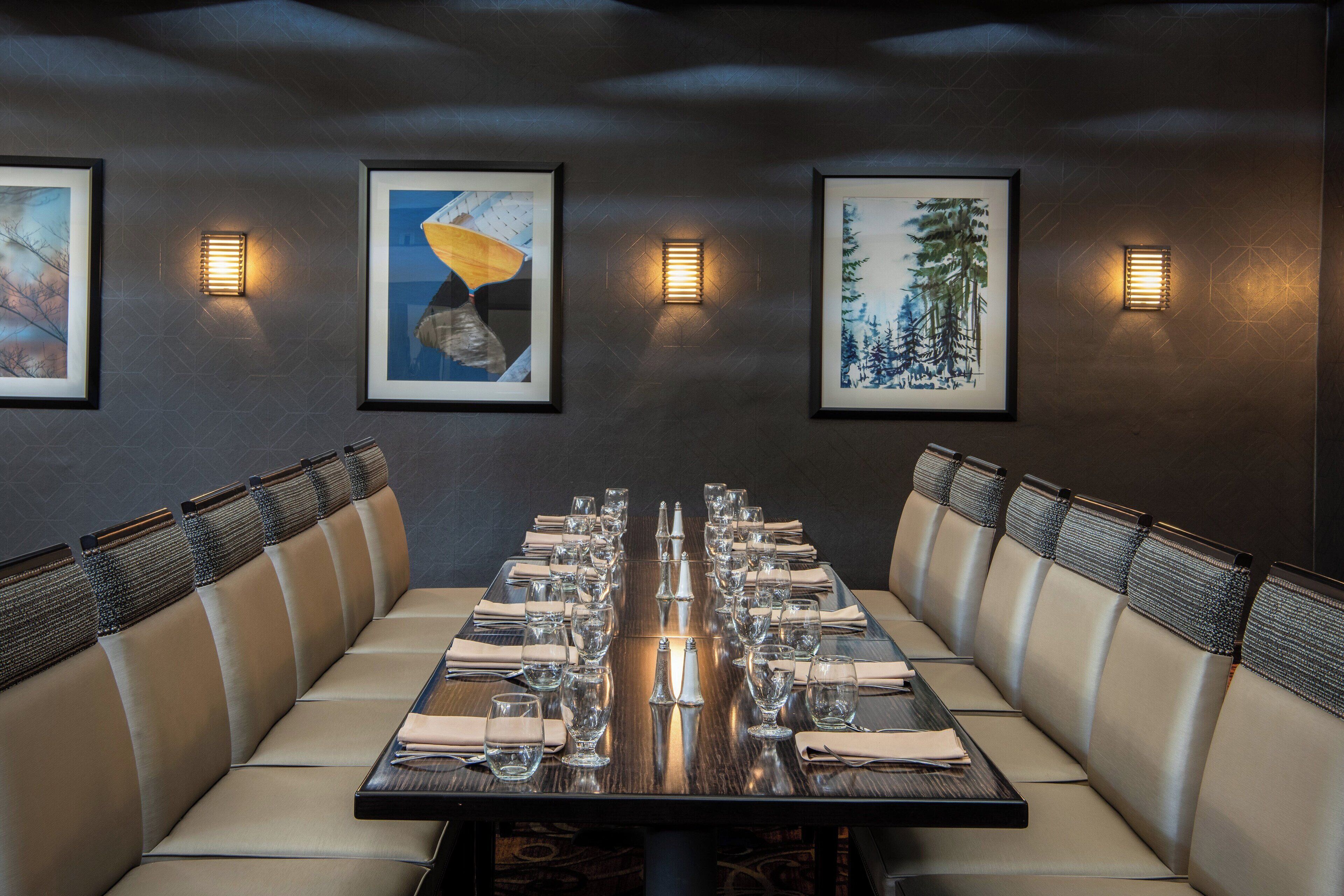 Una larga mesa de comedor con múltiples sillas, obras de arte enmarcadas en las paredes e iluminación instalada en la pared.