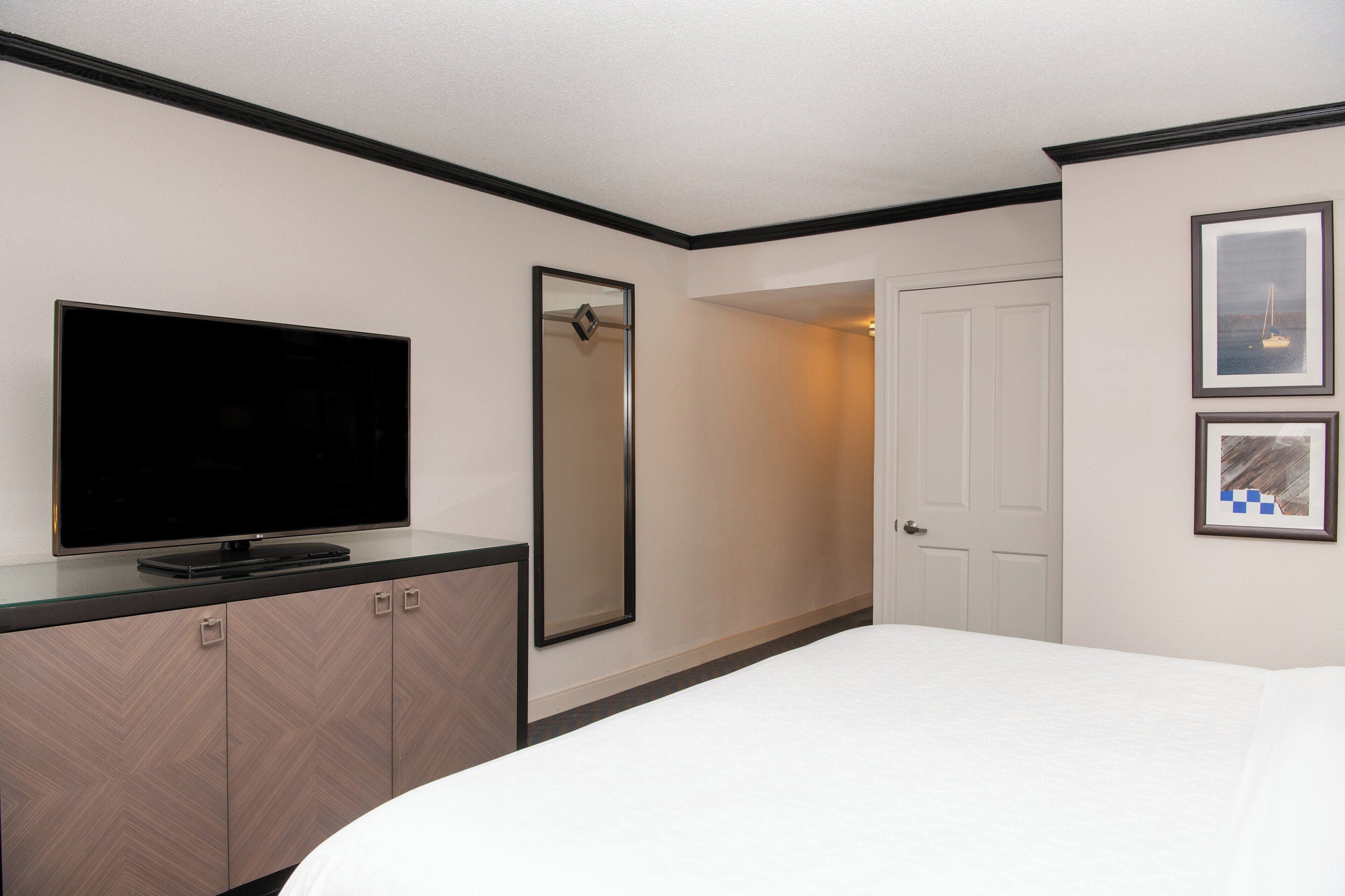 Una habitación de hotel con una cama, un televisor sobre un mueble, un espejo y dos cuadros en la pared.