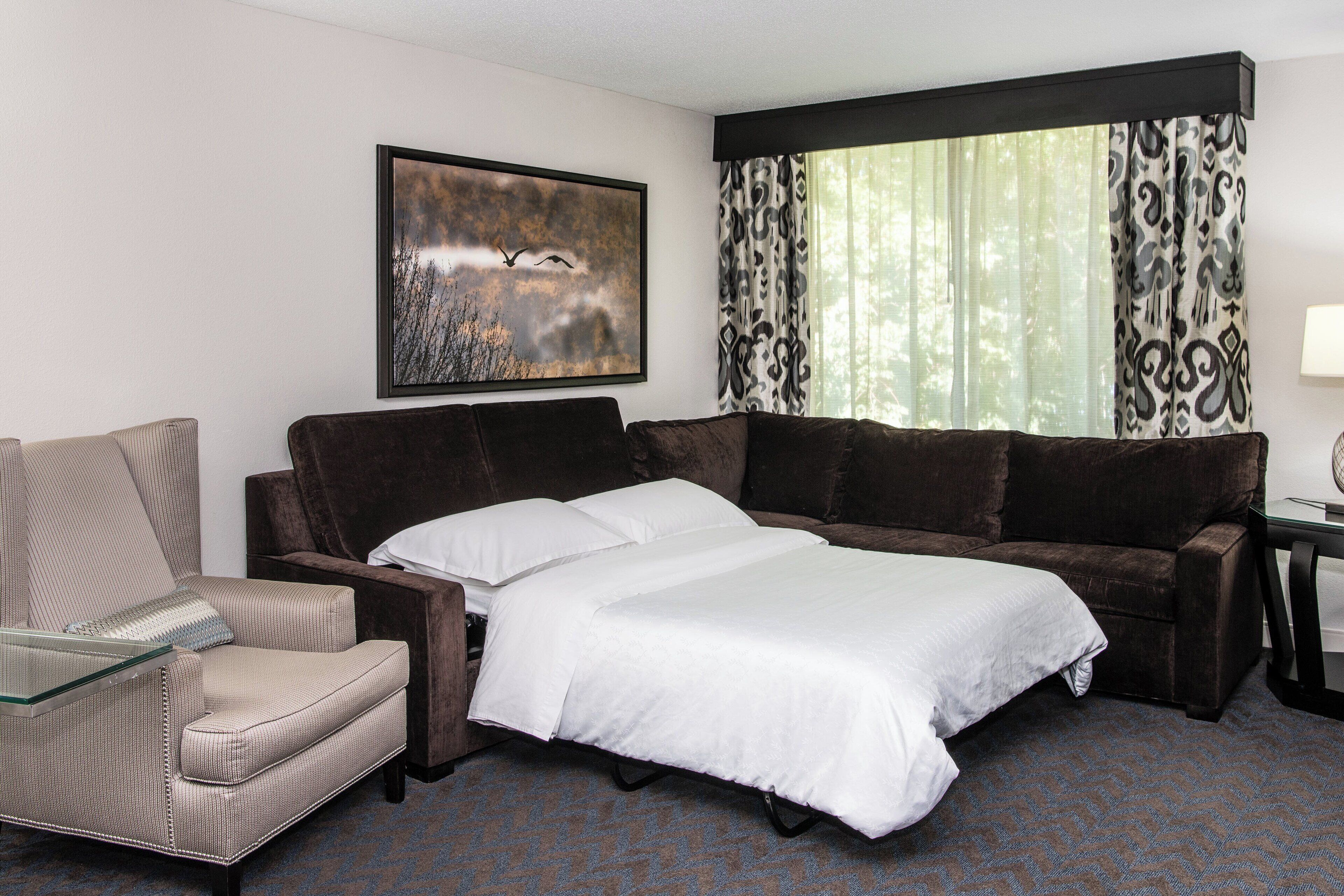 Une chambre d’hôtel avec un lit, un canapé, une chaise et une grande fenêtre ornée de rideaux à motifs.