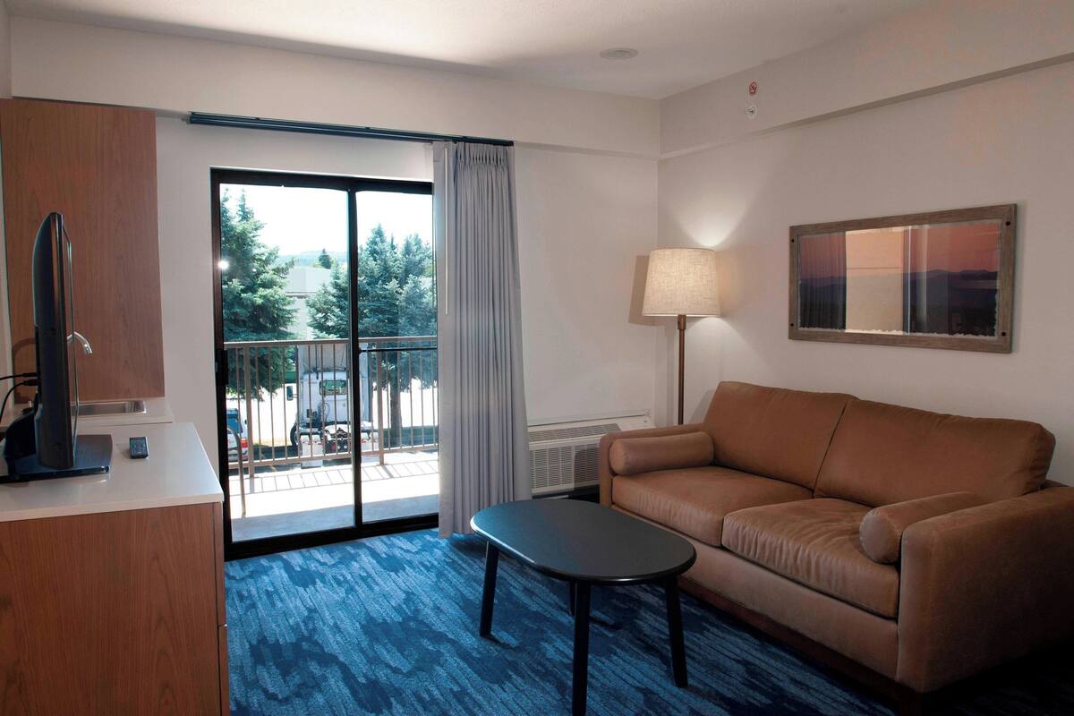 A hotel room with a brown leather sofa, a small black table, a TV on a wooden cabinet, a large window with a view of trees and a balcony, and a floor lamp.