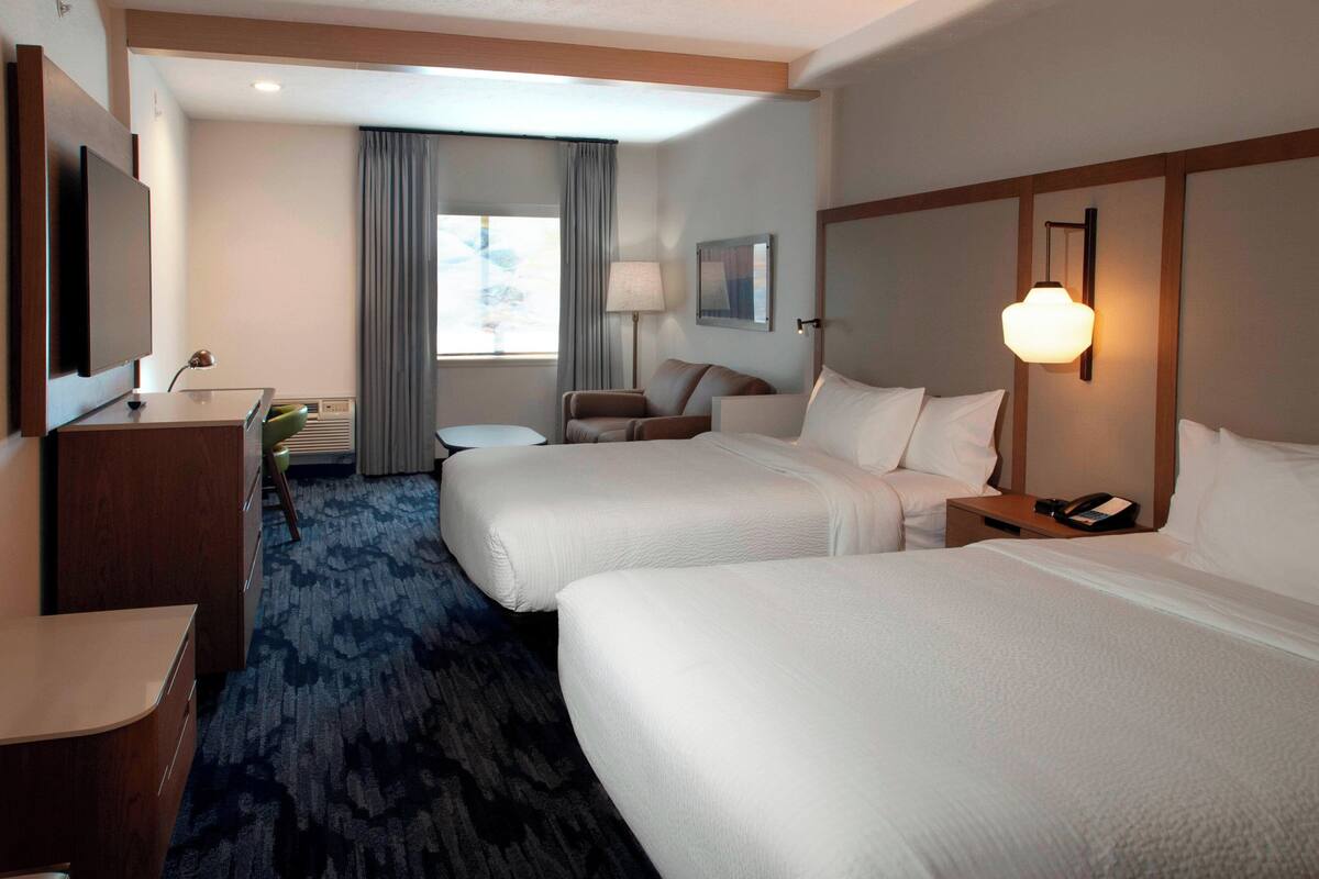 A hotel room with two beds, a desk, a chair, a lamp, and a window with curtains.