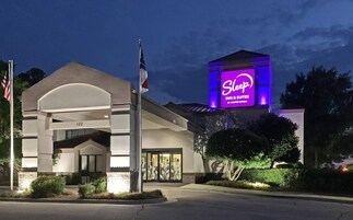 Edificio de Sleep Inn & Suites by Choice Hotels con un letrero iluminado llamativo, un asta con la bandera estadounidense y un área de entrada bien cuidada.